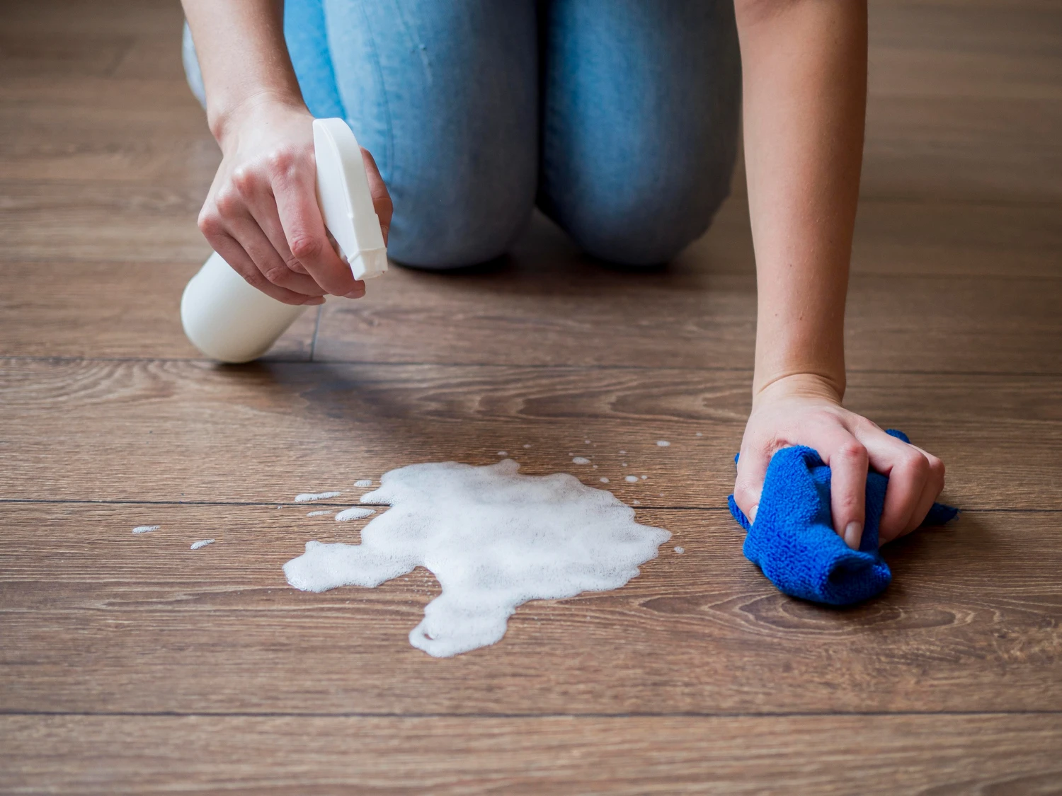 Person wischt hellen Vinylboden mit Sprayflasche und Tuch, entfernt sichtbaren Schaum.