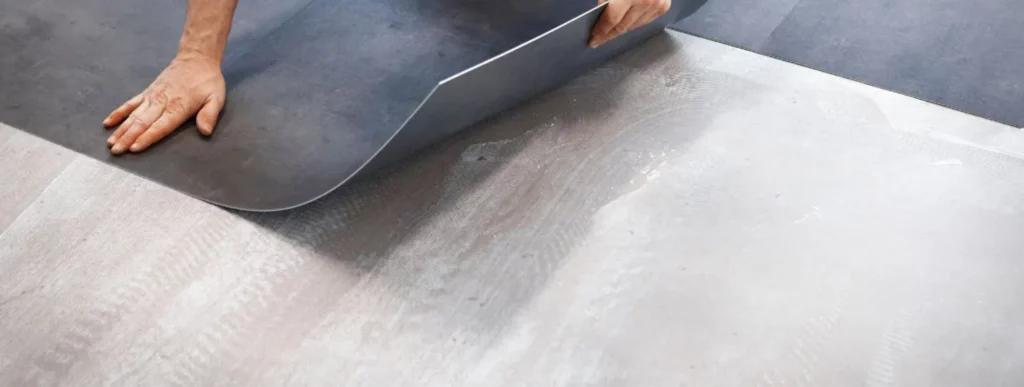 Zwei Hände arbeiten sorgfältig daran, ein dunkel gefärbtes Vinylblatt auf einem hellgrauen Betonboden zu legen. Der untere Teil des Vinyls ist leicht angehoben, wodurch die darunterliegende Oberfläche sichtbar wird. Dies deutet darauf hin, dass sich die Bodeninstallation oder Renovierung im Gange befindet. Die Hände sind entspannt und präzise positioniert, was ein geübtes Arbeiten vermuten lässt. Der Farbkontrast zwischen dem dunklen Vinyl und dem hellen Betonboden ist deutlich erkennbar und trägt zur klaren Struktur des Bildes bei. Die Szene vermittelt einen alltäglichen handwerklichen Prozess im Bereich Hausrenovierung oder Bodenpflege, passend zur Thematik von casando rund um Haus und Garten.
