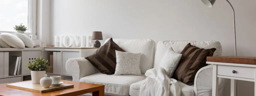Ein helles Wohnzimmer mit einem weißen Sofa, das mit gestreiften und strukturierten Kissen dekoriert ist. Ein Holztisch im Vordergrund trägt eine Topfpflanze und zwei weiße Tassen. Für Personen, die Einrichtungstipps für kleine Räume suchen, steht seitlich ein Schreibtisch mit einer Lampe. Auf einem Regal ist „HOME“-Dekoration platziert. Die Materialien sind überwiegend neutral und beruhigend gehalten, was dem Raum ein einladendes und gemütliches Ambiente verleiht.