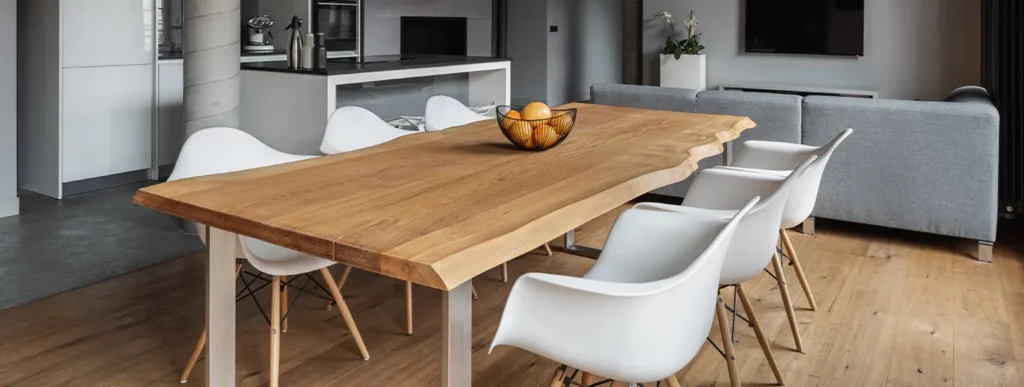 Ein gut beleuchteter, moderner Essbereich zeigt einen rechteckigen Holztisch in natürlichem Holzton, betont durch seine klare Maserung. Um den Tisch herum stehen sieben weiße Stühle mit schlichtem Design. Auf dem Tisch befindet sich eine transparente Schale, die mehrere rote Äpfel enthält und als dezente Dekoration dient. Im Hintergrund ist ein graues Sofa zu sehen, das Teil des offenen Raumkonzepts ist und sich zu einer Wand hin orientiert. An der Wand befindet sich ein Flachbildfernseher, der zum wohnlichen Ambiente montiert werden soll. Rechts vom Essbereich erstreckt sich eine funktionale Küche mit modernen Geräten und großzügiger Arbeitsfläche in neutralen Tönen, die nahtlos in den Raum integriert sind. Die gesamte Szenerie wirkt aufgeräumt und lädt zur gemeinsamen Nutzung von Wohn- und Essbereich ein.