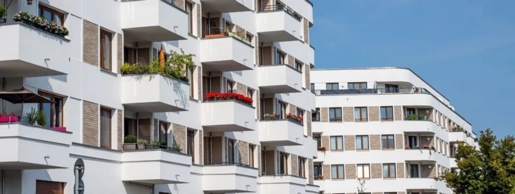 Das Bild zeigt moderne, weiße Mehrfamilienhäuser mit mehreren Balkonen, die Mietwohnungen beherbergen. Die Architektur zeichnet sich durch klare Linien und große Fenster aus. Einige Balkone sind mit Topfpflanzen und Blumen dekoriert, was den Charme des Gebäudes verstärkt. Im Hintergrund erstreckt sich ein klarer blauer Himmel, der die hellen Fassaden betont und in harmonischem Kontrast zur Architektur steht. Die Umgebung erweckt einen aufgeräumten und einladenden Eindruck, ideal für Wohnzwecke. Dieses Szenario könnte eine ruhige Wohnatmosphäre vermitteln sowie einen Bezug zu urbanem Wohnen schaffen.