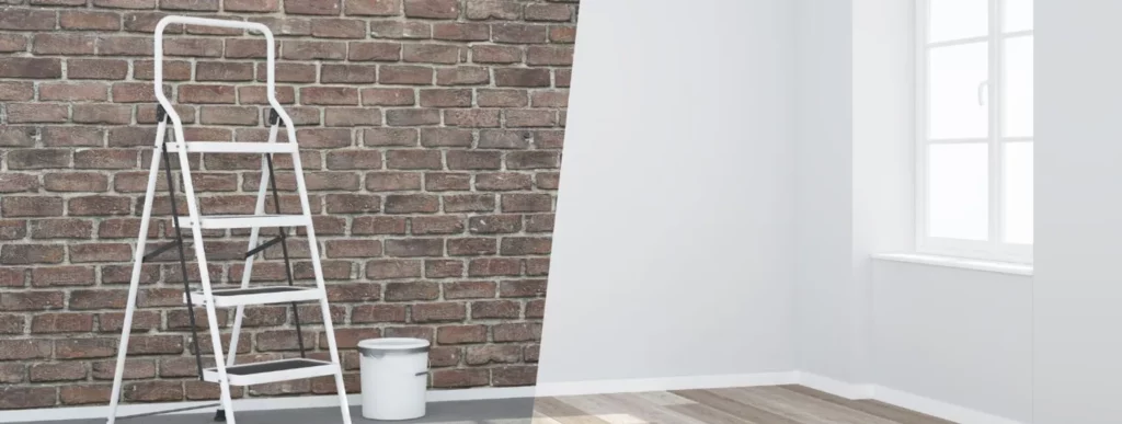 Das Bild zeigt einen Innenraum mit zwei unterschiedlichen Wandgestaltungen. Auf der linken Seite ist ein Zauberstab in Ziegeloptik zu sehen, während der rechte Zauberstab weiß gestrichen ist. Vor der Ziegelwand stehen ein weißer Leiter und ein Farbtopf, der auf anstehende Renovierungsarbeiten im Winter hindeutet. Rechts im Bild befindet sich ein Fenster, durch das Tageslicht den Raum erhellt. Der Boden besteht aus einem hellen Holzbelag, der den Raum freundlich wirken lässt.