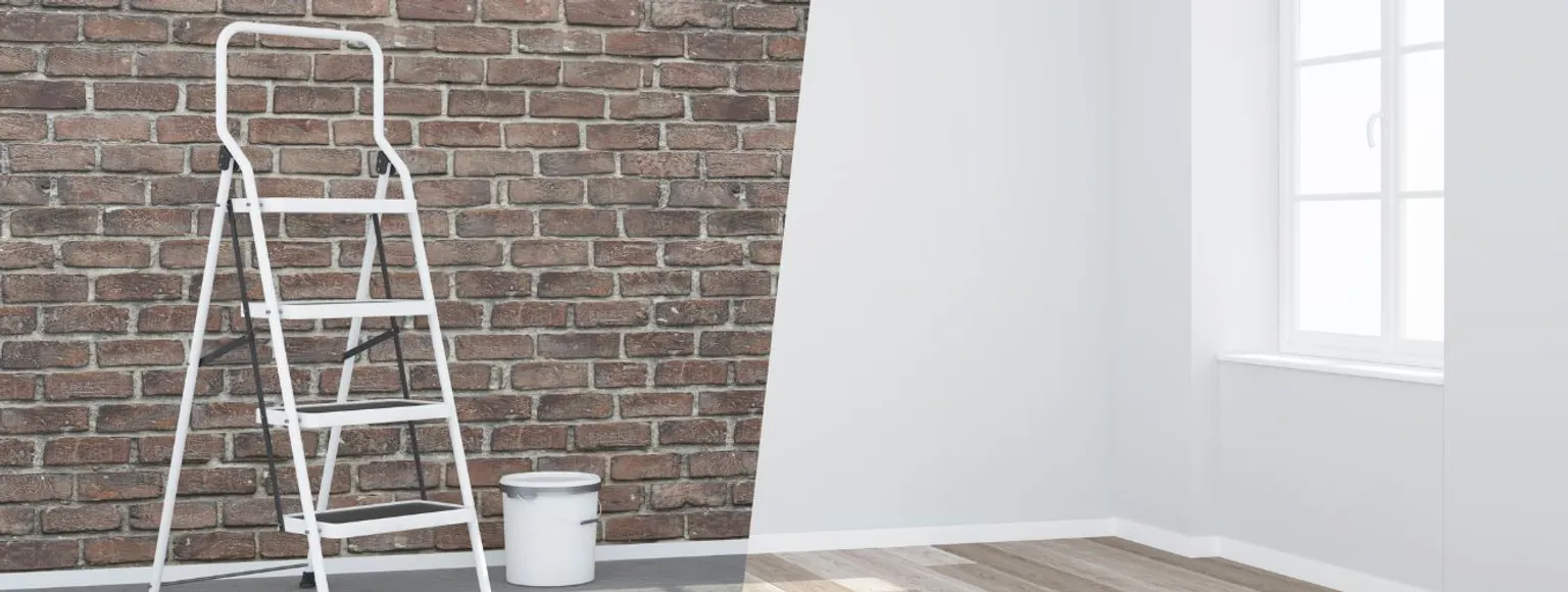 Das Bild zeigt einen Innenraum mit zwei unterschiedlichen Wandgestaltungen. Auf der linken Seite ist ein Zauberstab in Ziegeloptik zu sehen, während der rechte Zauberstab weiß gestrichen ist. Vor der Ziegelwand stehen ein weißer Leiter und ein Farbtopf, der auf anstehende Renovierungsarbeiten im Winter hindeutet. Rechts im Bild befindet sich ein Fenster, durch das Tageslicht den Raum erhellt. Der Boden besteht aus einem hellen Holzbelag, der den Raum freundlich wirken lässt.