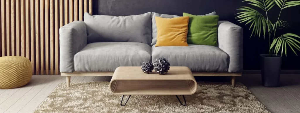 Das Bild zeigt einen modernen Wohnraum mit einem grauen Sofa, das mit orangefarbenen und grünen Kissen dekoriert ist. Vor dem Sofa steht ein hölzerner Couchtisch, der mit geometrischen Dekorationselementen geschmückt ist. Eine lebhafte grüne Topfpflanze verleiht dem Raum Frische. Der beige strukturierte Teppich auf dem Boden schafft Tiefe und trägt zur gemütlichen Atmosphäre bei. In der Ecke des Raumes befindet sich eine gelbe Fußstütze in Form eines Poufs, die farblich auf die Holzwandlamellen abgestimmt ist und harmonisch in das Gesamtbild integriert wird.