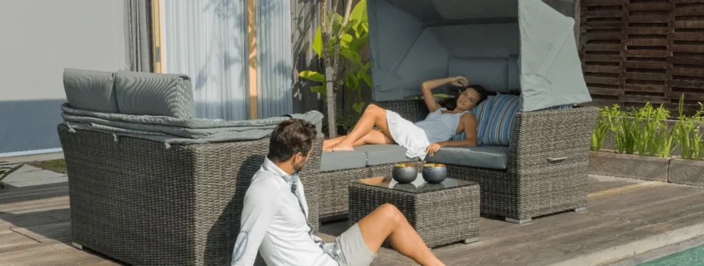 Eine Frau in einem weißen Kleid liegt entspannt auf einem grauen Outdoor-Sofa aus Rattan mit Überdachung. Das Sofa befindet sich auf einer Terrasse und bietet eine gemütliche Möglichkeit zur Einrichtung. In der Nähe sitzt ein Mann in einem hellen Hemd und Shorts entspannt auf dem Holzboden der Terrasse. Auf dem Sofa stehen zwei kleine Schalen, während im Hintergrund hohe Pflanzen entlang des modernen Gebäudes sichtbar sind. Die Szene vermittelt eine ruhige Atmosphäre von Entspannung und stilvoller Gestaltung im Außenbereich.