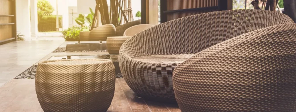 Die Abbildung zeigt eine einladende Outdoor-Sitzgruppe, die auf einer Holzterrasse platziert ist. Die Möbel bestehen aus Polyrattan und umfassen runde, geflochtene Tische sowie ein passendes Sofa. Das Sofa zeichnet sich durch seine geradlinige Form und neutrale Farbgebung aus. Umgeben wird die Szene von üppigem Grün, darunter verschiedene Pflanzenarten, die eine natürliche Begrenzung zur Umgebung bilden. Im Hintergrund ist teilweise eine Gebäudeaußenwand sichtbar. Das Tageslicht fällt weich auf die Sitzgruppe und verstärkt den natürlichen Charakter der Einrichtung. Diese Anordnung eignet sich gut für entspannte Stunden im Freien und verleiht dem Gartenbereich eine angenehme Atmosphäre.