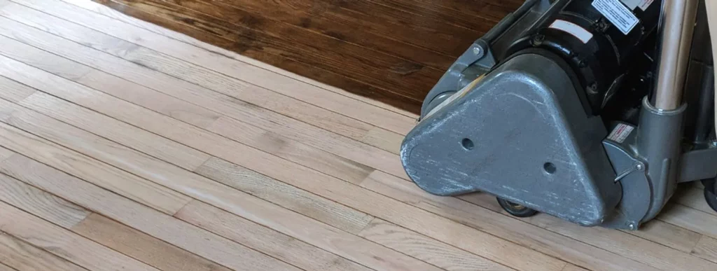 Ein Bild zeigt eine Fußbodenschleifmaschine, die auf einem zweifarbigen Holzboden im Einsatz ist. Auf der linken Seite des Bodens ist das Holz in einem hellen Farbton sichtbar, während die rechte Seite einen dunkleren, polierten Abschluss aufweist. Dies verdeutlicht den Fortschritt des Schleifvorgangs und das Potenzial für Oberflächenveränderungen beim Parkett abschleifen. Im Zentrum sieht man den trapezförmigen metallischen Basisteil der Schleifmaschine in Aktion, dessen Oberfläche bearbeitet und geglättet wird. Die Umgebung deutet darauf hin, dass es sich um eine Innenraumsituation ohne sichtbare Möbel oder Dekorationen handelt.