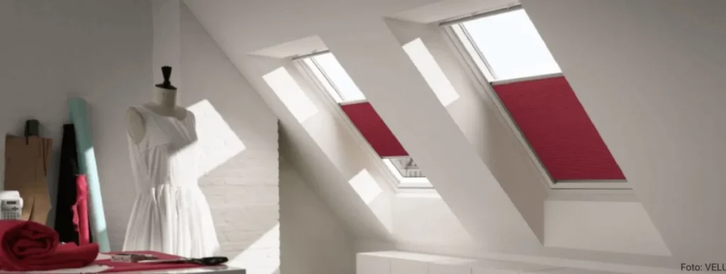 In einem Dachgeschossraum sind zwei Dachfenster mit roten Jalousien zu sehen, die Schatten auf die weißen Wände werfen. Links im Bild steht eine Schaufensterpuppe, die ein weißes Kleid trägt. Neben dieser befindet sich ein Tisch, auf dem rote und türkisfarbene Stoffrollen liegen. Die Umgebung vermittelt einen kreativen und organisierten Eindruck durch die klaren Farbakzente der Jalousien und Stoffe im Raum.