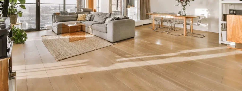 Das Bild zeigt einen geräumigen Wohn- und Essbereich, der von großen Fenstern dominiert wird. Im Vordergrund befindet sich ein graues Ecksofa, das auf einem texturierten Teppich steht. Vor dem Sofa ist ein gläserner Couchtisch platziert. Im Hintergrund steht ein hölzerner Esstisch mit sechs Stühlen, der eine gemütliche und einladende Atmosphäre schafft. Tageslicht strömt durch die Fenster und erzeugt Schattenmuster auf dem Laminatboden. Die Farbgebung des Raumes ist in sanften Erdtönen gehalten, was eine harmonische Umgebung unterstreicht.