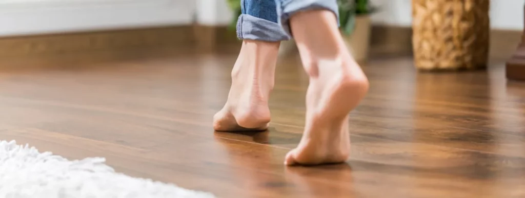 Eine Person geht barfuß über einen Holzboden aus Parkett. Die Hosenbeine der Person sind hochgekrempelt, was auf eine entspannte und informelle Umgebung hindeutet. Neben dem Fuß ist ein weißer Teppich zu sehen, der sanft den Boden bedeckt. Im Hintergrund ist ein geflochtener Korb unscharf erkennbar. Dies alles deutet auf eine gemütliche Innenraumatmosphäre hin, möglicherweise verstärkt durch die Wärme einer Fußbodenheizung im Raum. Die Farben der Szene sind überwiegend warm und neutral, was den Eindruck einer behaglichen Wohnatmosphäre verstärkt.