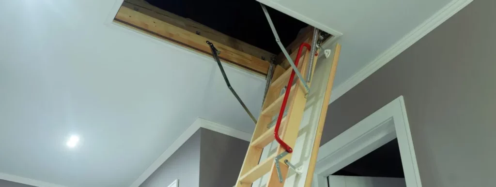 Das Bild zeigt eine ausgeklappte Bodentreppe, die von einer offenen Deckenluke in einen Raum mit grauen Wänden und weißen Zierleisten herabführt. Die Treppe ist mit Metallbeschlägen versehen und besitzt einen roten Griff, der beim Hinauf- oder Hinabsteigen als Haltemöglichkeit dient. Der Raum ist gut beleuchtet, wodurch die schräge Deckenkonstruktion klar zur Geltung kommt. Es sind keine Personen im Bild zu sehen; Der Fokus liegt auf der Funktionalität und dem Design der Bodentreppe, sowie auf den architektonischen Details des Raumes, wie den Kontrasten zwischen Wandfarbe und Leisten.