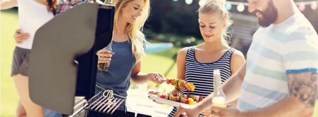 Drei Personen grillen Spieße auf einem Terrassengrill, die Sonne und die Sitzgelegenheiten im Freien sind sichtbar.