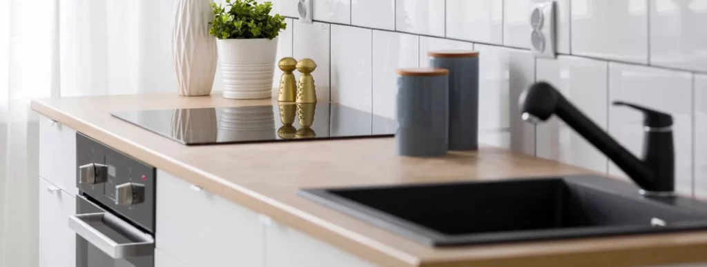 Das Bild zeigt eine moderne Küche mit einer Arbeitsplatte aus hellem Holz. Auf der Arbeitsfläche befinden sich ein schwarzes Elektrokochfeld und eine Spüle. Die Wand hinter der Küchenzeile ist mit weißen Fliesen verkleidet, die als Rückwand dienen. Neben dem Kochfeld sind zwei graue Behälter zu sehen, sowie eine Pflanze in einem weißen Blumentopf. Der Ofen, der über zwei Bedienknöpfe verfügt, ist in die Unterseite der Theke eingebaut. Die Umgebung wirkt ordentlich und funktional gestaltet und vermittelt einen effizienten Arbeitsplatz für kulinarische Tätigkeiten im Rahmen von Haus- und Gartenprojekten.