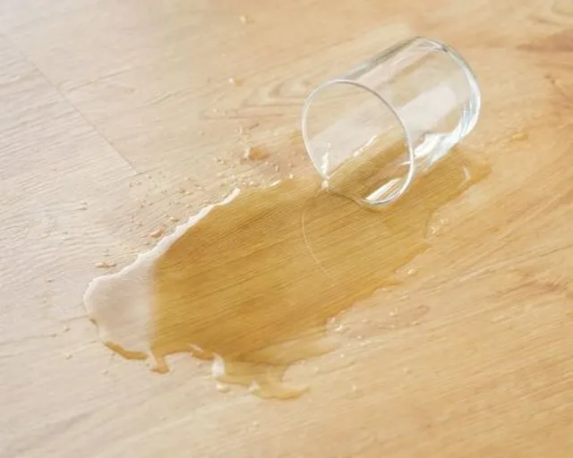 Ein durchsichtiges Glas liegt umgekippt auf dem hellbraunen Boden, um das herum Flüssigkeit verschüttet wurde.