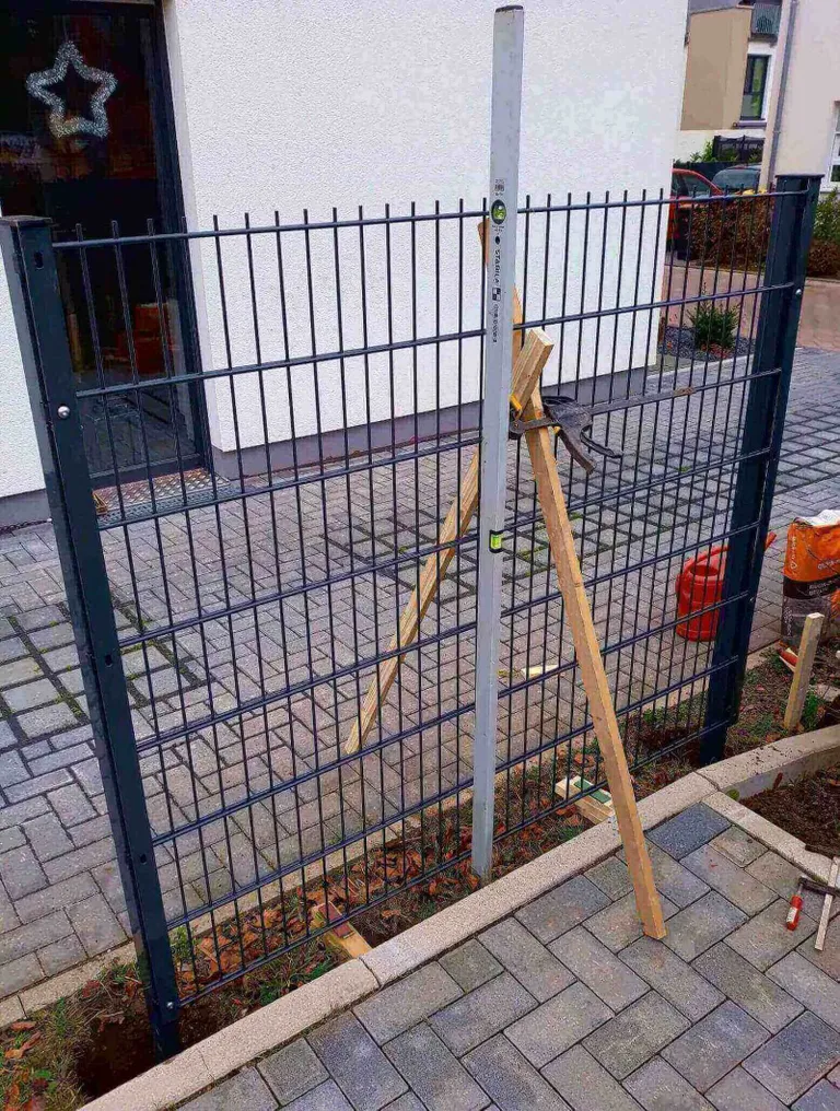 Ein grauer Doppelstabmattenzaun wird montiert, wobei Holzbretter als Stütze dienen.
