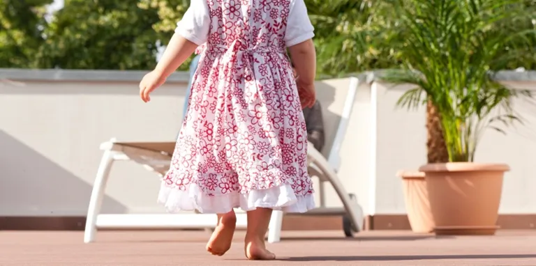 Ein Kind ohne Schuhe in einem hellen Kleid läuft auf einer grauen WPC-Terrasse neben einer grünen Topfpflanze.