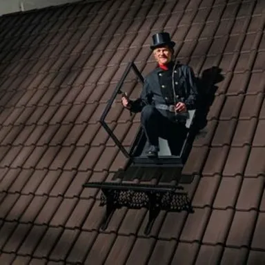 Ein Schornsteinfeger in schwarzer Uniform mit Zylinder sitzt neben einer Dachfenster-Austauschluke.