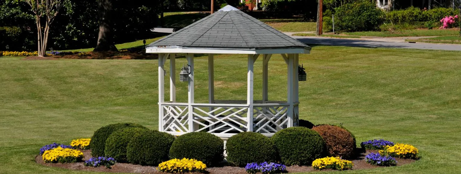 Ein weißer Gartenpavillon steht auf grünem Rasen, umgeben von Büschen und bunten Blumen.
