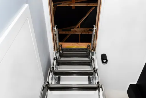 Eine hölzerne Dachbodenleiter, die von einer weißen Deckenluke heruntergezogen wird und zu einem dunklen Dachbodenraum führt.