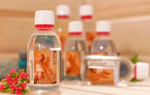 Eine klare Flasche mit Saunaaufguss vor anderen Flaschen und Blumen auf einer hellen Fläche.