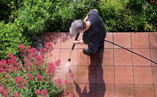 Eine Person, die rote Terrassenfliesen mit einem Hochdruckreiniger säubert, in der Nähe blühende rosa Blumen im Freien.
