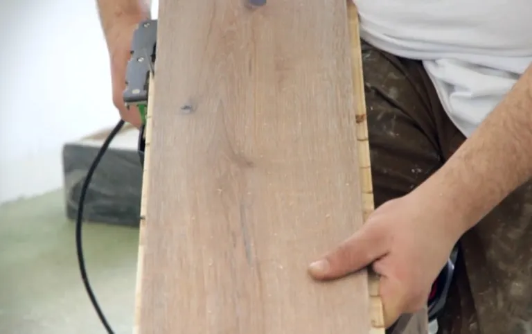 Eine Person hält eine hellbraune Holzdiele und ein Elektrowerkzeug in der Hand, während sie die Bodenverlegung vorbereitet.