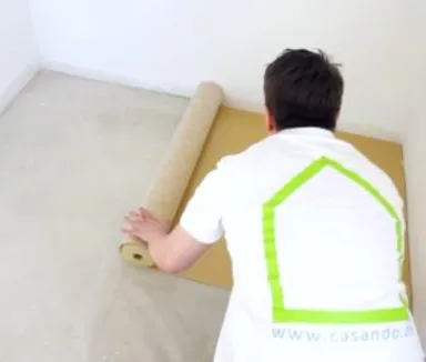 Eine Person legt eine Laminat-Unterlage auf hellem Boden in einem leeren Raum aus.