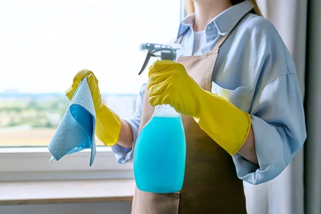 Eine Person mit Handschuhen hält eine Sprühflasche in der Nähe des Fensters und bereitet sich auf die Reinigung und Abdichtung der Türkanten vor.
