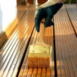 Eine Person mit Handschuhen trägt im Freien mit einem Pinsel Holzbeize auf eine braune Terrassenterrasse auf.