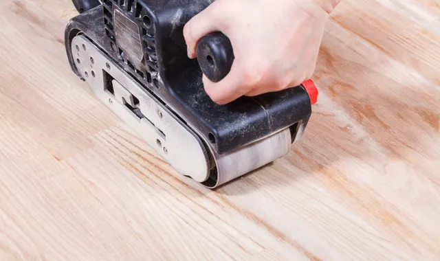 Eine Person schleift einen hellen Holzboden in einer Mietwohnung mit einer Bandschleifmaschine.