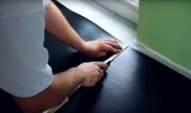 Eine Person schneidet einen schwarzen Laminatboden in der Nähe einer grünen Wand für die Verlegung einer Fußbodenheizung zu.