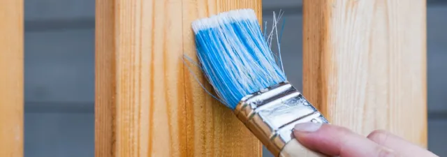 Eine Person streicht mit einem Pinsel mit blauem Stiel die Holzplanken eines Gartenhauses braun.