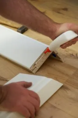 Eine Person trägt vor dem Zusammenbau eines Glastürrahmens Klebstoff auf leichte Holzverkleidungsteile auf.