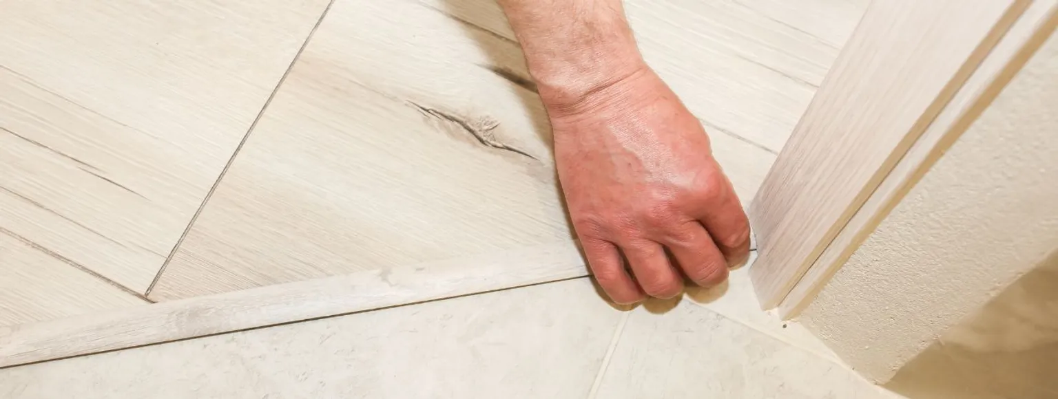 Eine Person verlegt ein Übergangsprofil aus Holz zwischen zwei hellen Bodenbelägen im Innenbereich.