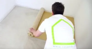 Eine Person verlegt einen hellbraunen Vinylboden in der Ecke eines leeren, farblich neutralen Raums.