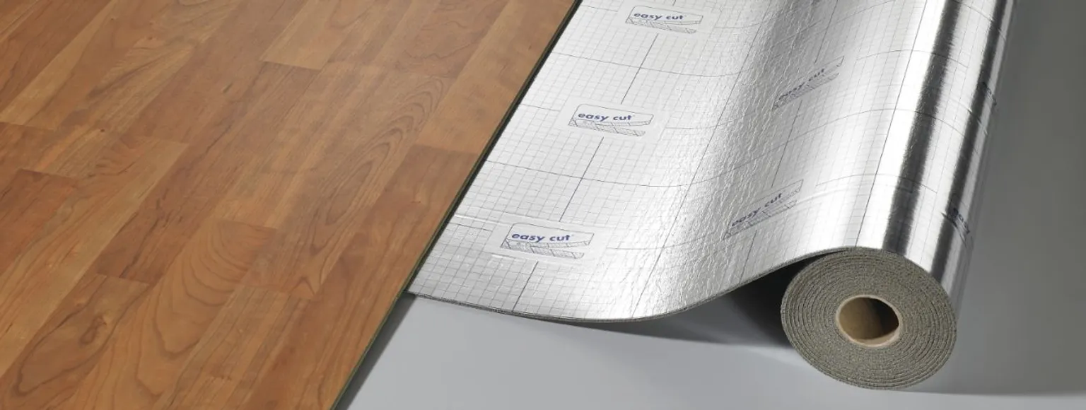 Eine Rolle Akustikunterlage wird während der Verlegung neben den hellbraunen Laminatboden gelegt.
