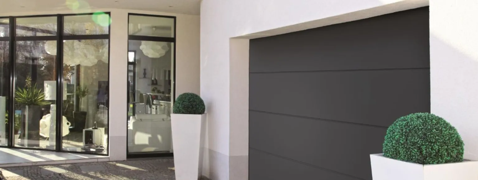 Eingang eines modernen Hauses mit großen Glasfenstern, Garagentor und Grünpflanzen im Topf.