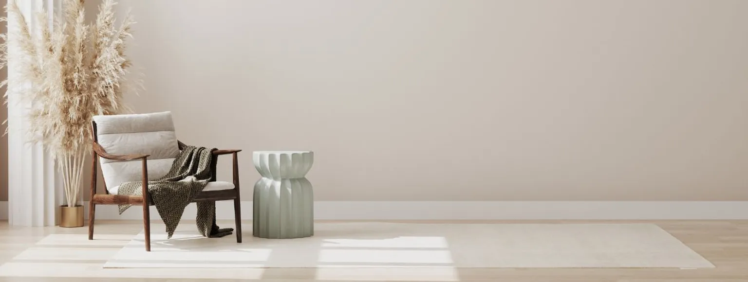 In einem minimalistischen Raum ist ein Holzstuhl mit einem beigen Kissen und einer dunkelgrünen Decke zu sehen. Der Stuhl steht auf hellbraunen Vinylböden. Neben dem Stuhl befindet sich ein zylinderförmiger, hellgrüner Beistelltisch. In der Ecke des Raumes steht ein hoher Strauß getrockneter Pampasgrasstängel. Die Wände und die Dekorationsgegenstände des Raumes sind in beruhigenden neutralen Tönen gehalten, was eine harmonische Atmosphäre schafft.
