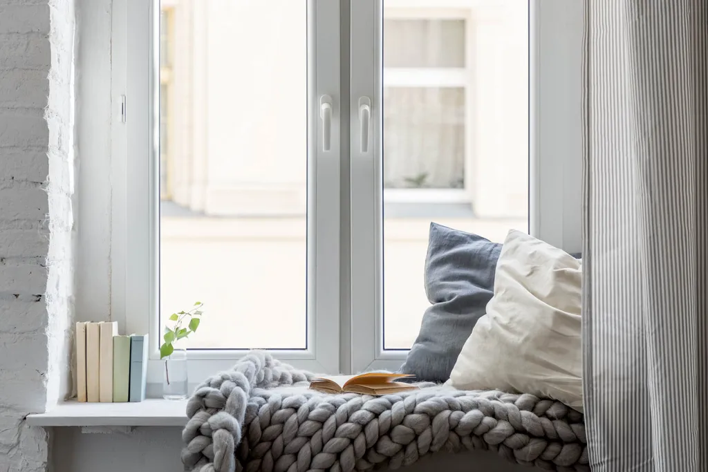 Fensterbank mit grauen Kissen, beiger Decke, Bücherstapel, Grünpflanze am Holzfenster.