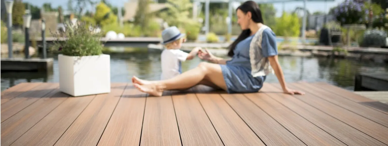 Frau und Kind sitzen auf einem grauen WPC-Deck am Wasser, umgeben von der Natur.