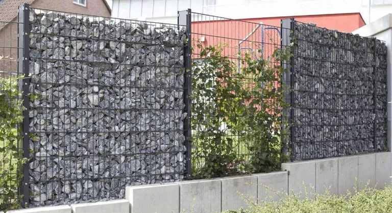 Gabionendrahtzäune, gefüllt mit grauen Steinen, gesäumt von grünen Blattpflanzen und Sträuchern.