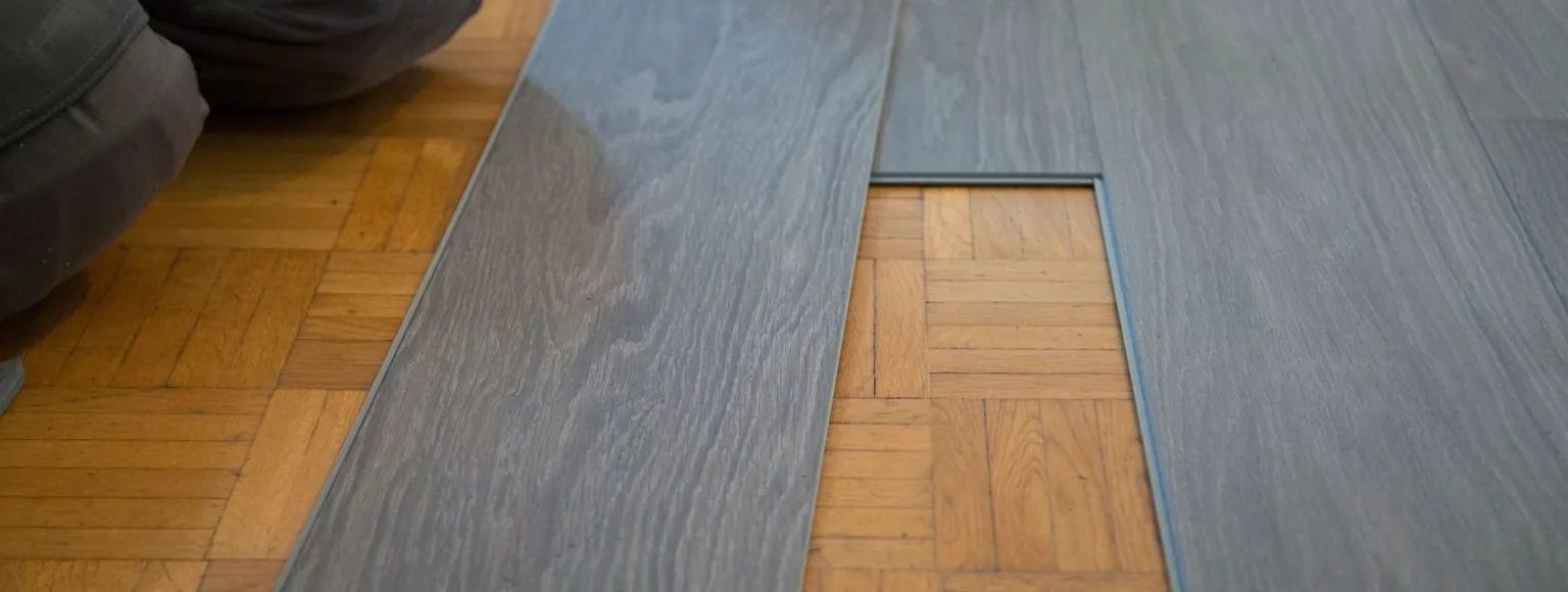 Graue Laminatdielen werden bei der Verlegung über einen braunen Parkettboden gelegt.