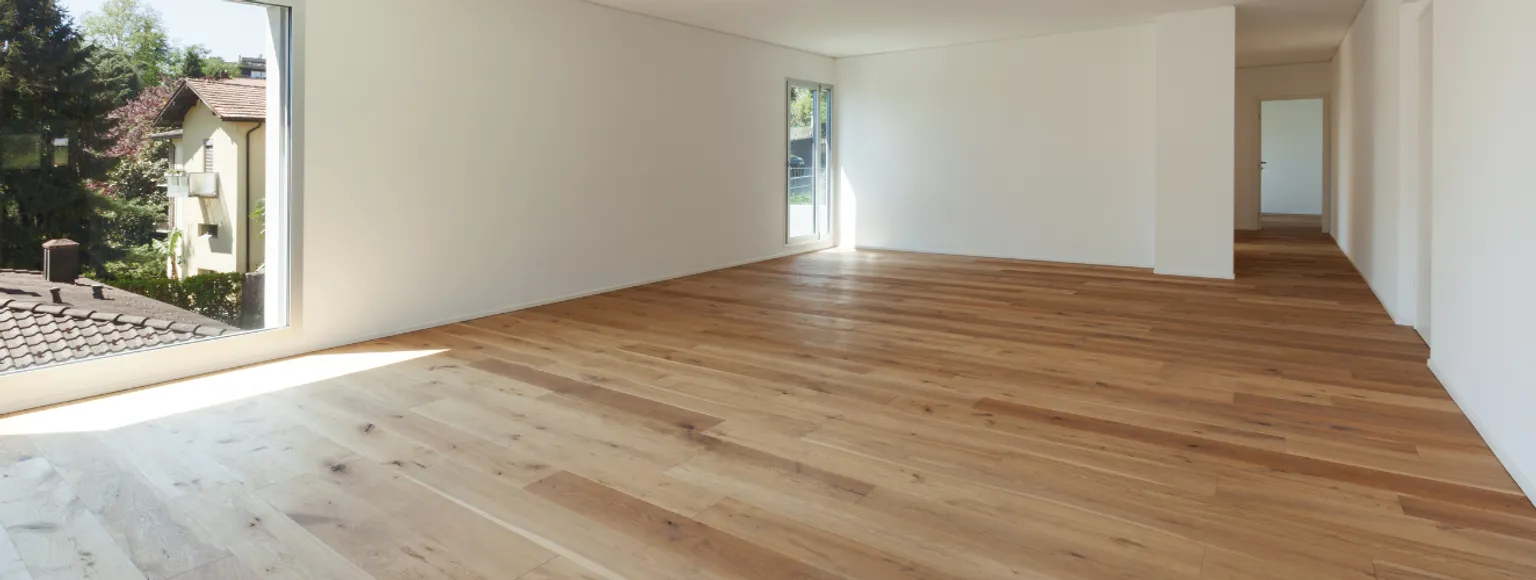Helles Zimmer mit weißen Wänden, großen Fenstern und ordentlich verlegten Parkettstäben.