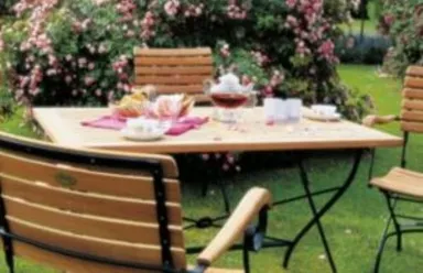 Holztisch mit Teeservice und Gebäck, umgeben von Metallgartenmöbeln und Blumen.