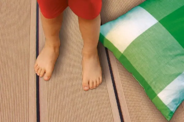 Kind mit nackten Füßen, das auf einer gestreiften Matte in der Nähe einer Terrassenüberdachung und eines Kissens steht.