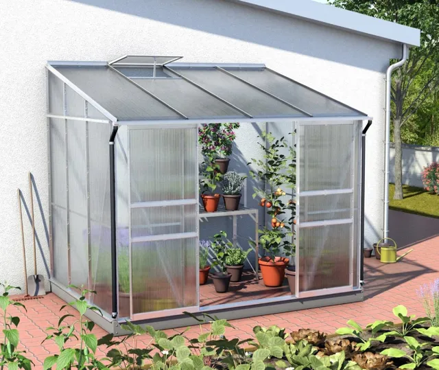 Kompaktes Gewächshaus aus Glas und Metall in kleinem Garten neben Holzzaun und Terrasse.