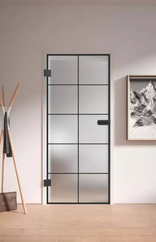Mattierte Lofttür mit schwarzem Gitterrahmen, neben einem Garderobenständer und Wandkunst an einer hellen Wand.