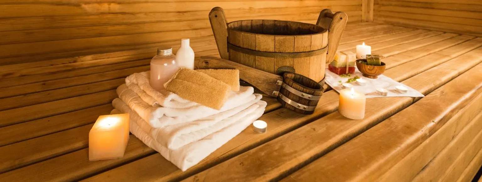 Mehrere gefaltete Handtücher, weiße Kerzen und Wellnessartikel auf einer Holzfläche in der Nähe einer Sauna.