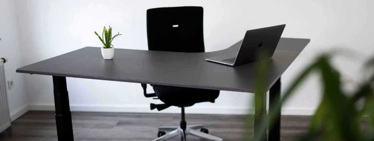 Moderner höhenverstellbarer Schreibtisch mit grauem Bürostuhl, Laptop und grüner Pflanze.