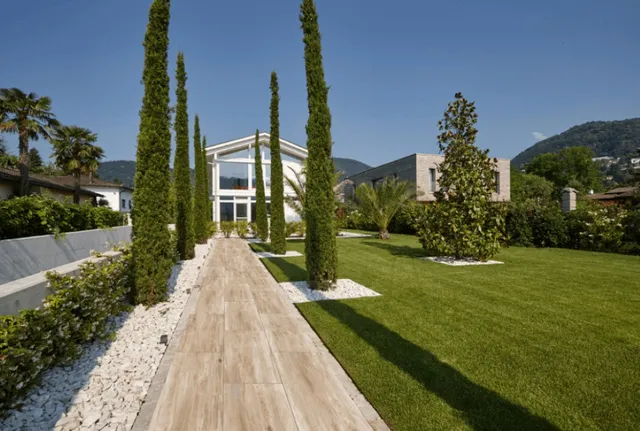 Modernes Haus mit Gehweg aus Steinplatten, grünem Rasen und hohen Bäumen im Gartenbereich.